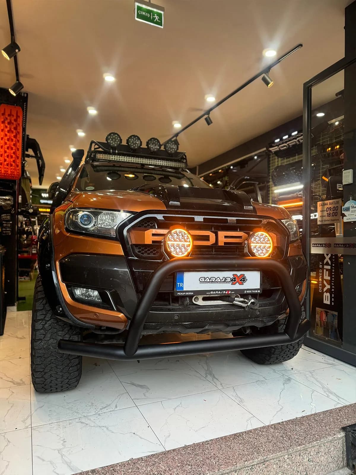 Mağazanın içinde turuncu Ford Ranger: tavan LED barı, yuvarlak spotlar, FORD harfli panjur, ön koruma demiri ve vinç
